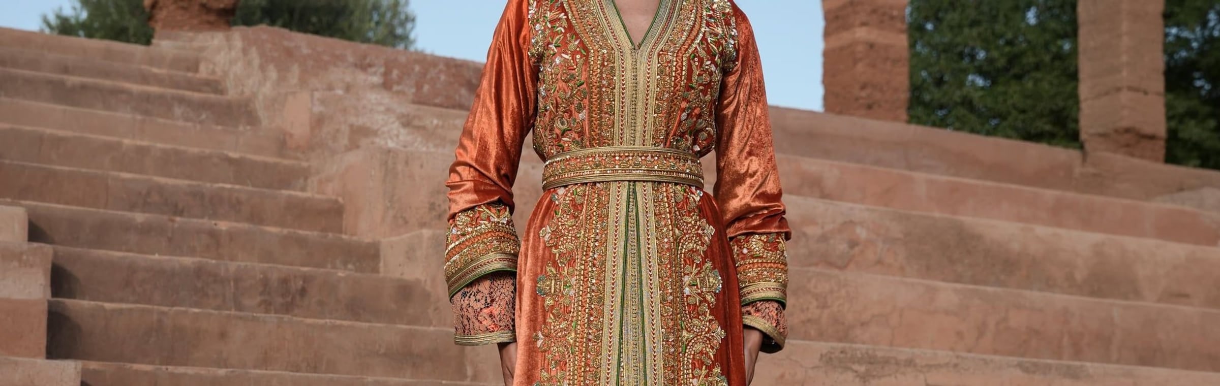 Rust velvet Moroccan kaftan with gold and green embroidery against warm stone