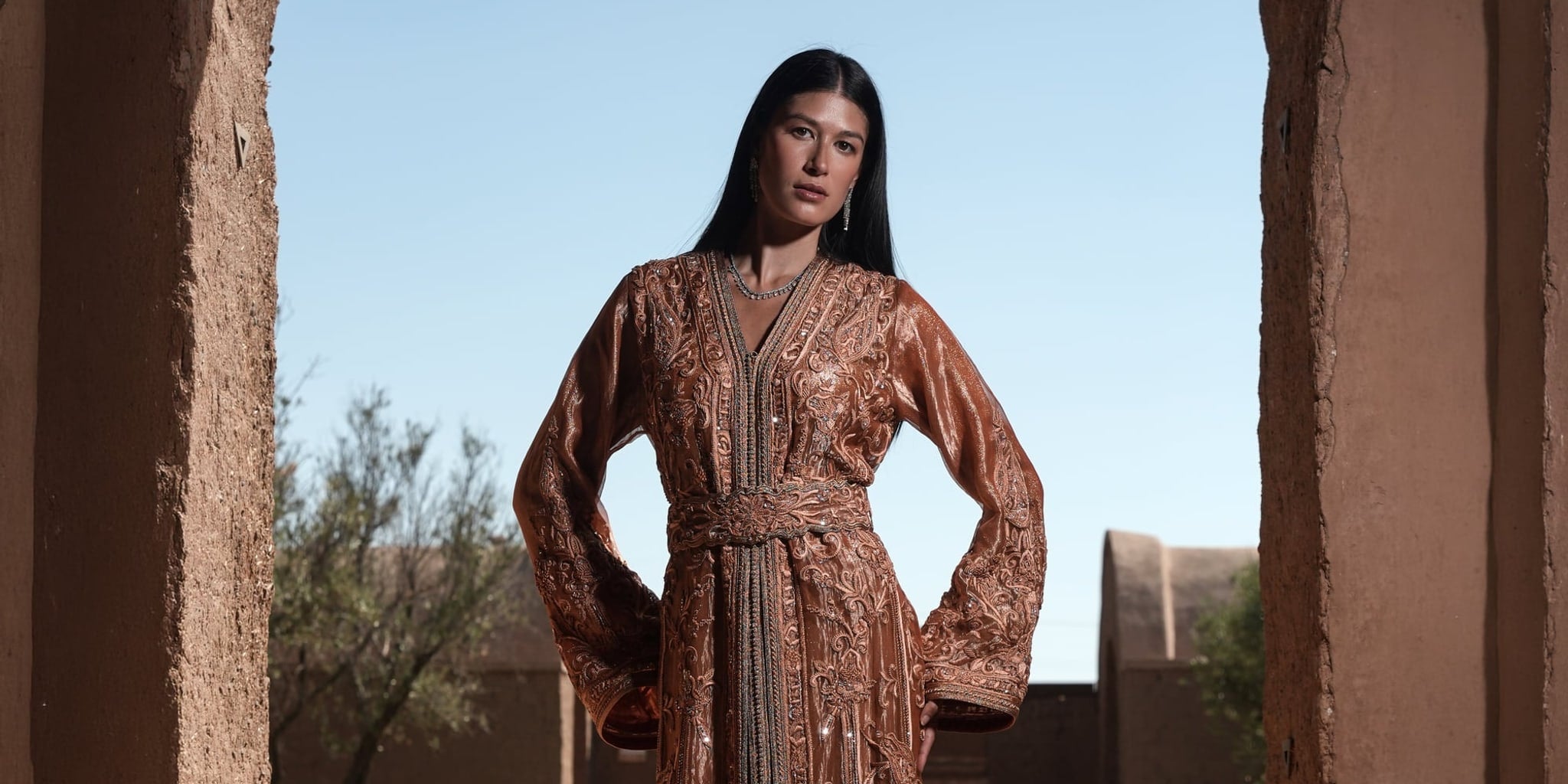 Copper Solstice bronze embroidered Moroccan kaftan framed by an adobe archway